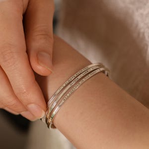 May include: Three silver-colored, engraved bangles stacked on a wrist. The top bangle reads "IT'S ALWAYS BEEN YOU", the middle one says "OLIVIA, LIAM", and the bottom one says "MAY FOREVER IN OUR HEARTS". The bangles are simple and elegant.