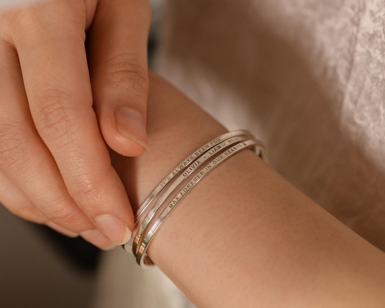 May include: Three silver-colored, engraved bangles stacked on a wrist. The top bangle reads "IT'S ALWAYS BEEN YOU", the middle one says "OLIVIA, LIAM", and the bottom one says "MAY FOREVER IN OUR HEARTS". The bangles are simple and elegant.