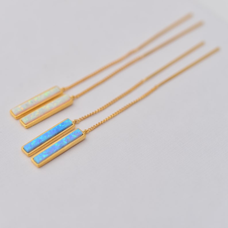 May include: Two pairs of gold-tone threader earrings featuring rectangular bar pendants. One pair contains white iridescent opal-style inlays, while the other pair features bright blue iridescent opal-style inlays set in polished gold metal.