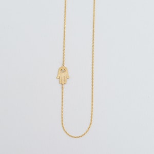Könnte beinhalten: Eine goldene Kettenkette mit einem Hamsa-Hand-Anhänger mit einem kleinen Diamanten in der Mitte.