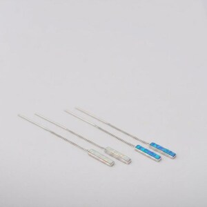 May include: A pair of silver-toned threader earrings featuring long, thin chains attached to rectangular bar pendants. One pendant displays a shimmering white iridescent inlay, while the other pendant features a vibrant blue iridescent inlay.