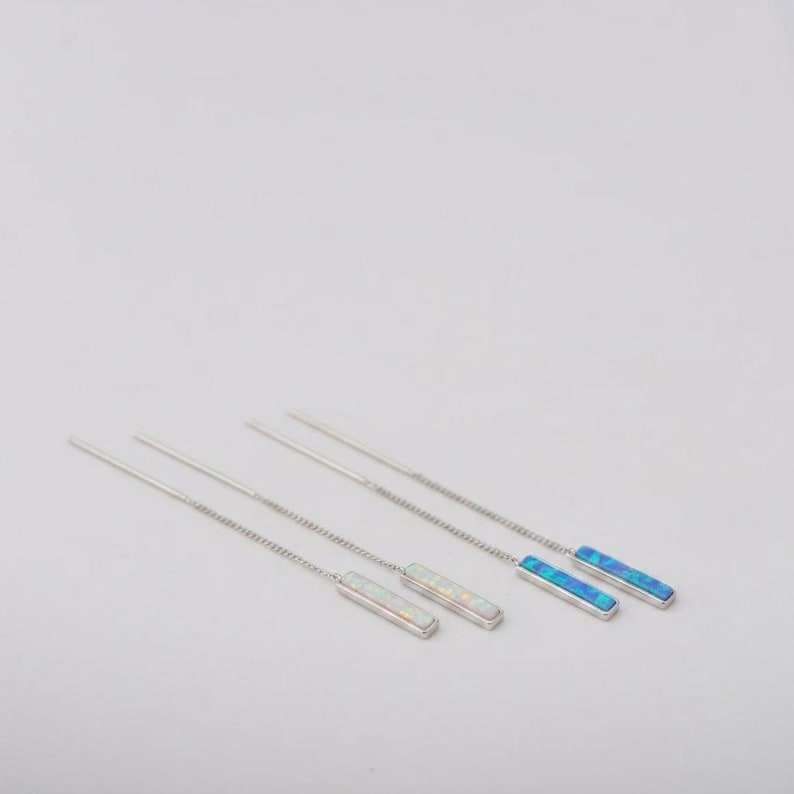 May include: A pair of silver-toned threader earrings featuring long, thin chains attached to rectangular bar pendants. One pendant displays a shimmering white iridescent inlay, while the other pendant features a vibrant blue iridescent inlay.