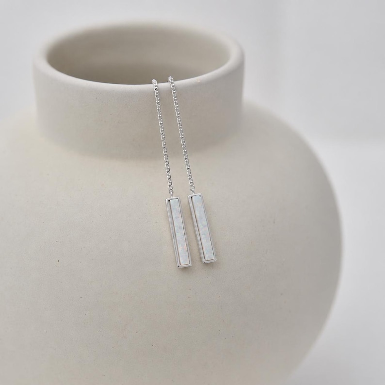 May include: A pair of silver-toned threader earrings featuring long, slender chains that connect to rectangular bar pendants. Each pendant has a smooth, metallic frame surrounding a flat, white, rectangular inlay with a subtle, pearlescent finish.
