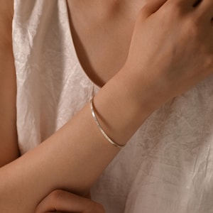 May include: A silver bangle bracelet with engraved text. The bracelet is worn on a person's arm, with a white top in the background. The bracelet is a simple, elegant piece of jewelry.