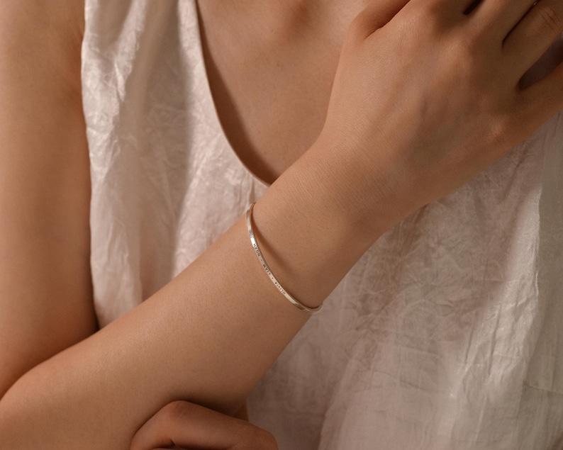 May include: A silver bangle bracelet with engraved text. The bracelet is worn on a person's arm, with a white top in the background. The bracelet is a simple, elegant piece of jewelry.
