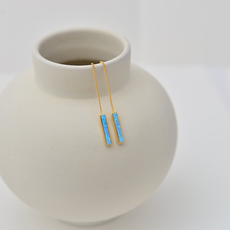 May include: A pair of gold-tone threader earrings featuring long, slender rectangular bars inlaid with vibrant, iridescent blue opal-like stones. The earrings consist of delicate gold chains that connect to the vertical bar pendants.