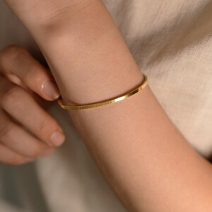 May include: A gold-colored, thin bangle bracelet. The bracelet is worn on an arm and has engraved text. The arm is against a neutral-colored background. The bracelet is a simple, elegant piece of jewelry.