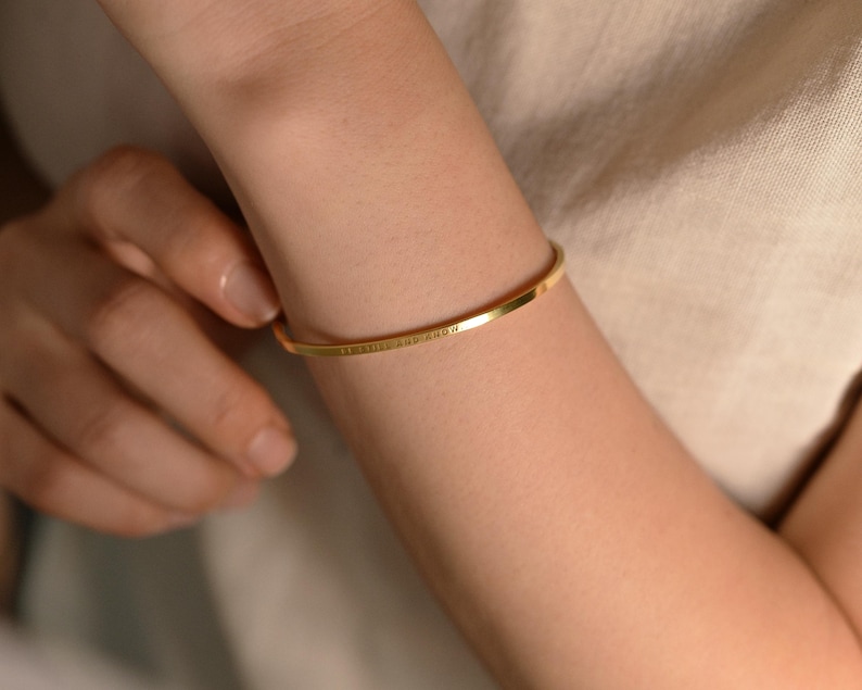 May include: A gold-colored, thin bangle bracelet. The bracelet is worn on an arm and has engraved text. The arm is against a neutral-colored background. The bracelet is a simple, elegant piece of jewelry.