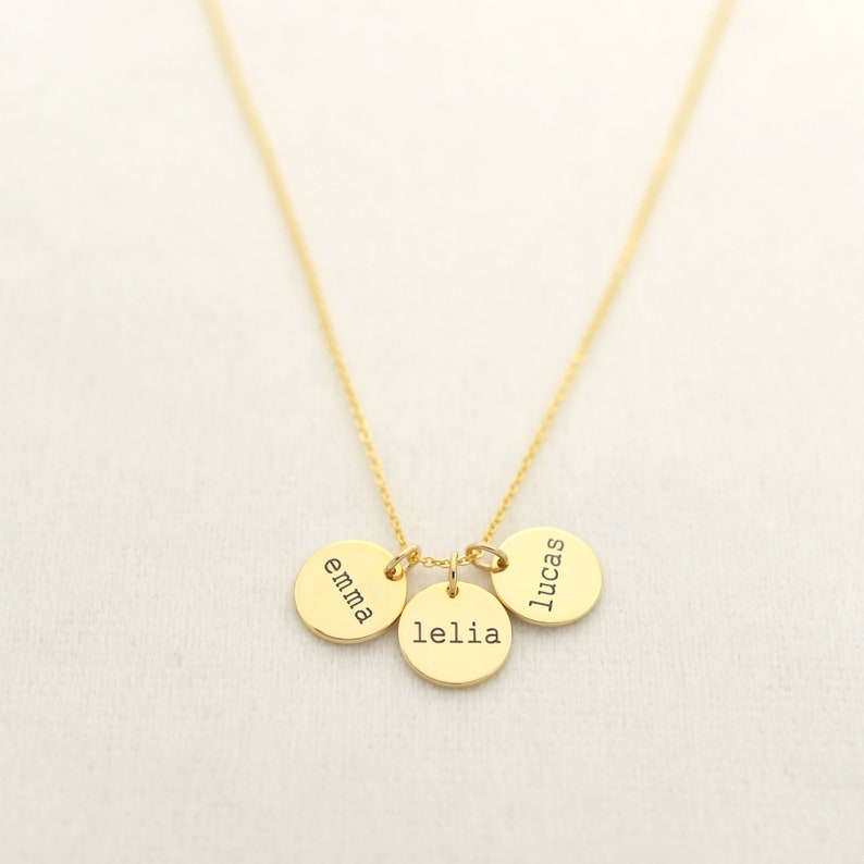 Op de afbeelding: Gouden ketting met drie ronde bedels, elk gegraveerd met een naam: Emma, Lelia en Lucas.