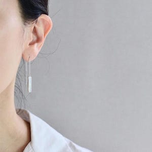 May include: A silver threader earring featuring a slender, rectangular pendant with a white, iridescent inlay. The earring consists of a thin chain that passes through the earlobe, ending in a straight metal bar on one side and the pendant on the other.