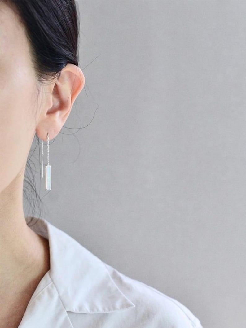 May include: A silver threader earring featuring a slender, rectangular pendant with a white, iridescent inlay. The earring consists of a thin chain that passes through the earlobe, ending in a straight metal bar on one side and the pendant on the other.