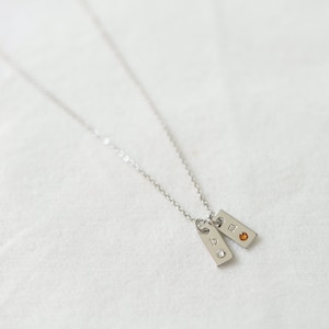 Op de afbeelding: Een zilveren ketting met twee kleine rechthoekige hangers. De bovenste hanger heeft een kleine oranje edelsteen en de onderste hanger heeft een kleine heldere edelsteen. De hangers zijn gegraveerd met de letters "19" en "90".