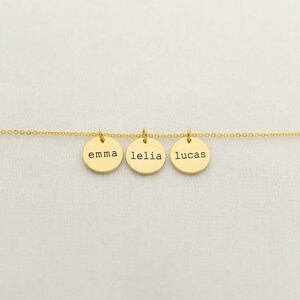 Op de afbeelding: Gouden ketting met drie ronde bedels, elk gegraveerd met een naam: Emma, Lelia en Lucas.