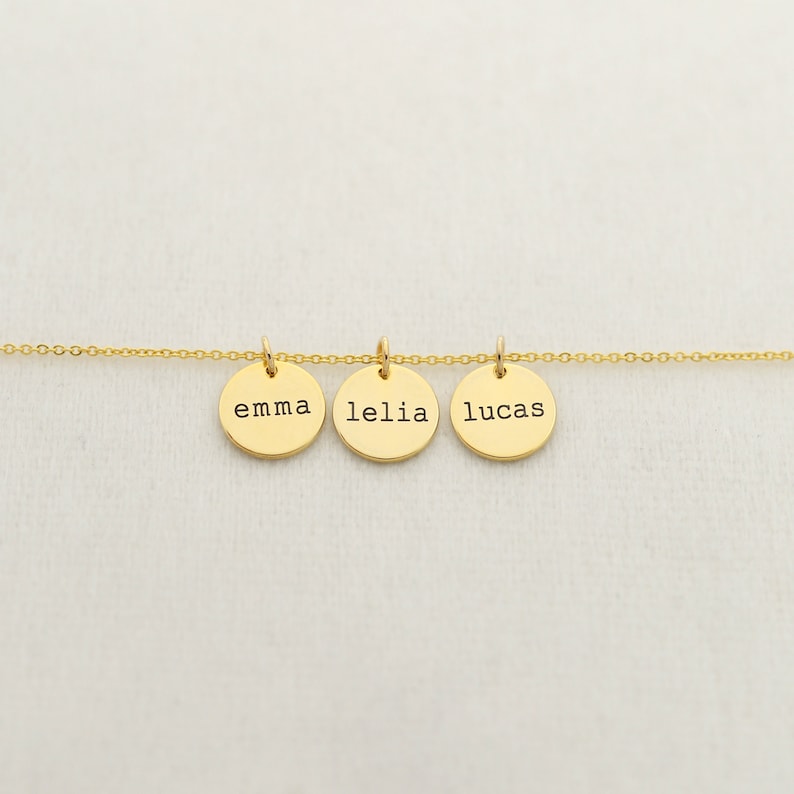Op de afbeelding: Gouden ketting met drie ronde bedels, elk gegraveerd met een naam: Emma, Lelia en Lucas.