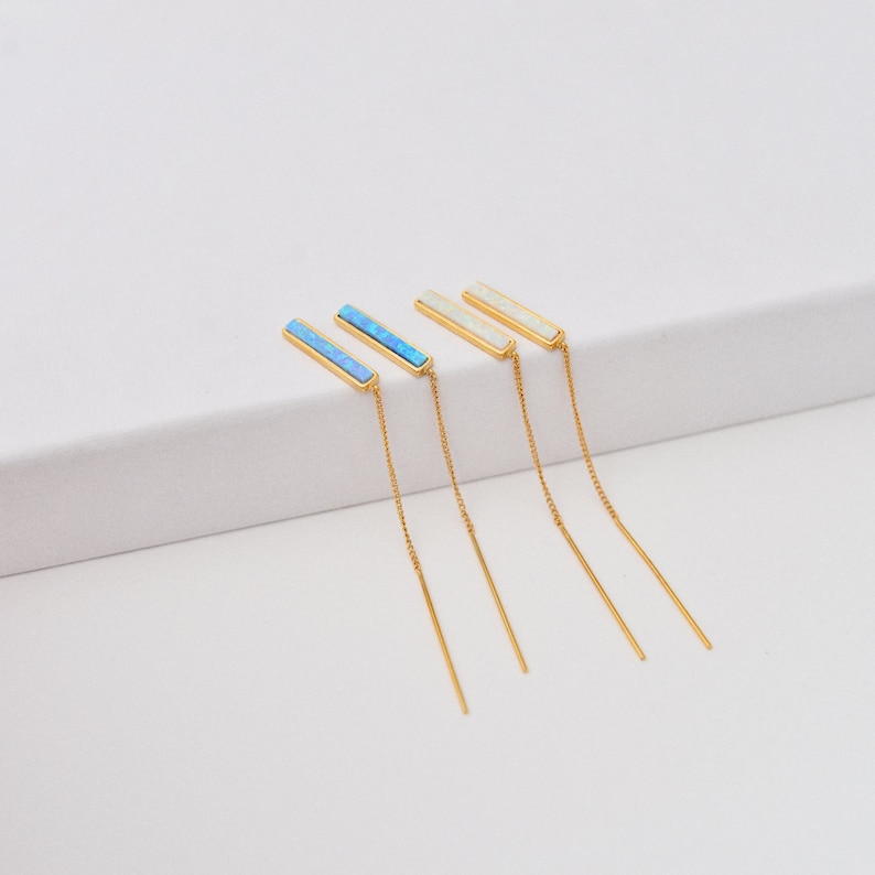 May include: Two pairs of gold-tone threader earrings. One pair features rectangular bars with bright blue inlay, while the other pair features rectangular bars with white inlay. Each earring consists of a thin gold chain attached to the bar.