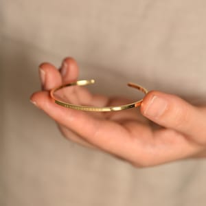 May include: A gold-colored, open cuff bracelet with the inscription "LET IT BE AND KNOW". The bracelet is held in a hand against a neutral background. The bracelet is a simple, elegant design.