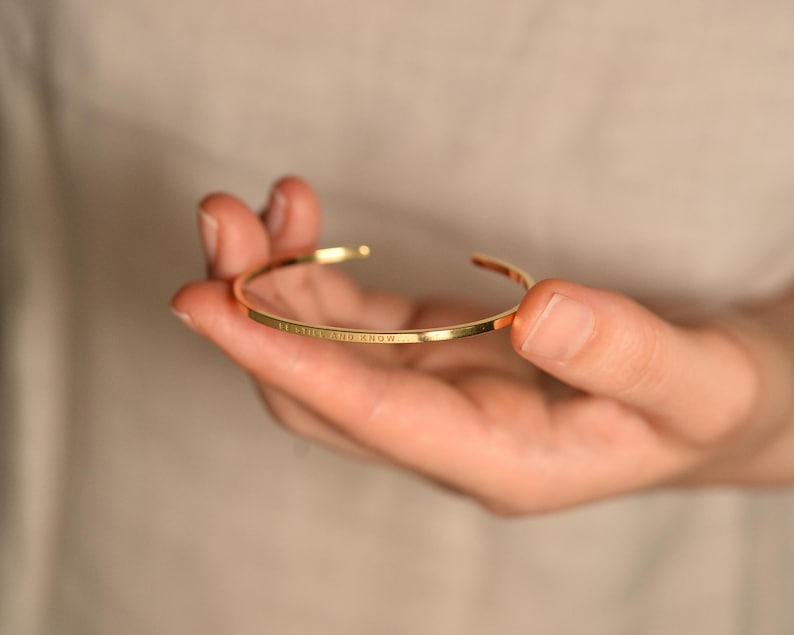 May include: A gold-colored, open cuff bracelet with the inscription "LET IT BE AND KNOW". The bracelet is held in a hand against a neutral background. The bracelet is a simple, elegant design.