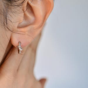 Puede incluir: Un pendiente de nudo de plata con un diseño sencillo. El pendiente se lleva en la oreja izquierda de una persona.