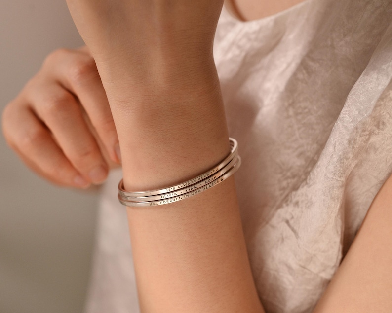 May include: Three silver-colored, engraved bracelets stacked on a wrist. The bracelets have text that reads "IT'S ALWAYS BEEN YOU", "OLIVIA + LIAM + NOAH", and "WALK FOREVER IN OUR HEARTS". The bracelets are worn on a person's arm.