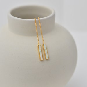May include: A pair of gold-tone threader earrings featuring long, slender rectangular bars. Each bar is inlaid with a white, iridescent opal-like stone that displays subtle flashes of color. The earrings hang from delicate, thin gold-tone chains.