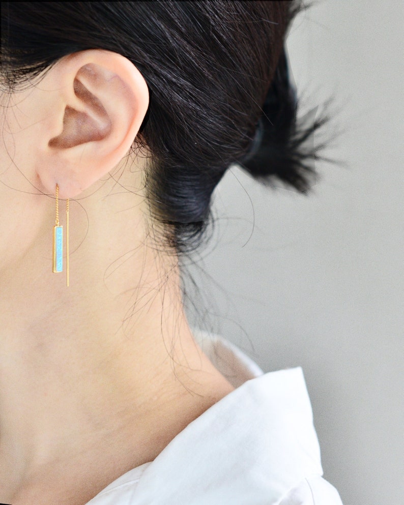 May include: A gold threader earring featuring a thin, vertical rectangular pendant inlaid with a bright turquoise stone. The earring consists of a delicate gold chain that passes through the earlobe, ending in a straight, slender gold post.