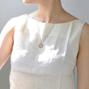 May include: A woman wearing a white sleeveless top with a silver chain necklace and a small gold pendant.