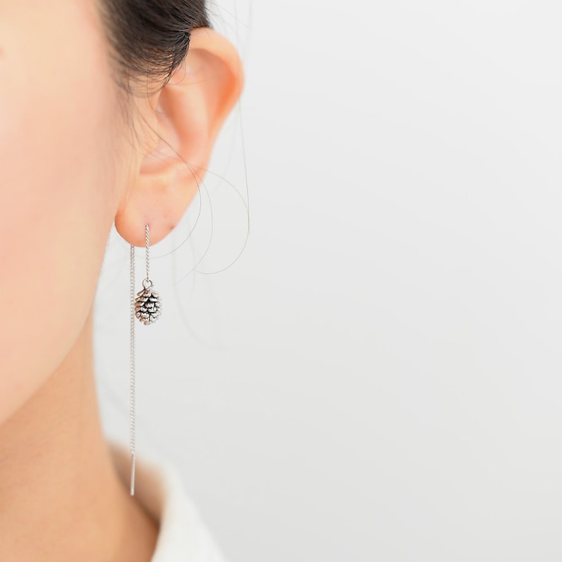 May include: Silver dangle earrings with a small pine cone charm hanging from a thin chain.