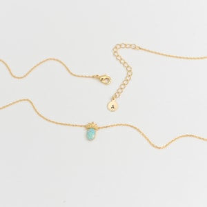 May include: A gold chain necklace with a small pineapple charm featuring a light blue opal stone. The necklace has a lobster clasp and an extender chain with a small round tag that says "A".