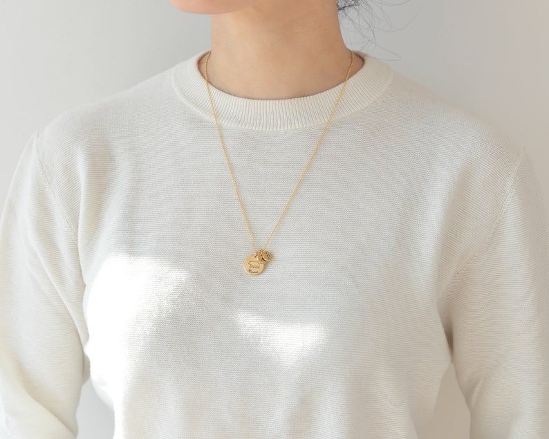 May include: A gold chain necklace with a round pendant that says "Stay Wild" and a smaller charm. The necklace is worn by a person wearing a white sweater.