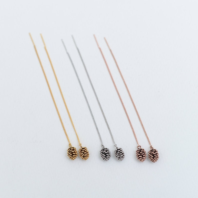 May include: Four pairs of delicate chain earrings with a small pine cone charm at the end. The earrings are available in gold, silver, and rose gold.