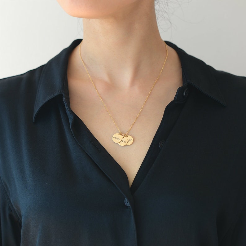 Op de afbeelding: Een gouden ketting met drie ronde hangers. Elke hanger heeft een ander nummer gegraveerd. De ketting wordt gedragen door een persoon met een zwart shirt.
