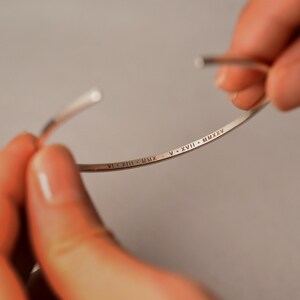 May include: A silver-colored bangle bracelet with engraved Roman numerals. The bracelet is open, held by two hands. The engraved text includes the Roman numerals VI XIII MMX and V XVII MMXXV.