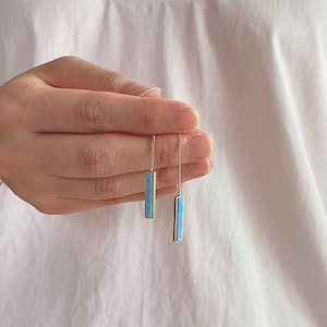 Puede incluir: Un par de pendientes de barra dorados con una incrustación azul claro similar al ópalo. Los pendientes cuelgan de delicadas cadenas doradas. Los pendientes se sostienen frente a una camisa blanca.