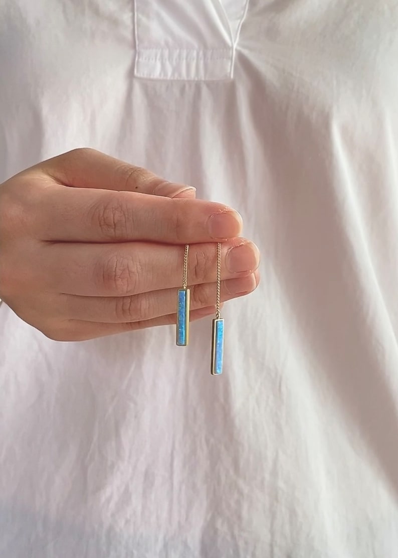 May include: A pair of gold-tone drop earrings featuring thin chains attached to rectangular pendants. Each pendant is inlaid with a vibrant, light blue iridescent stone that reflects light, creating a sleek and elongated vertical design.