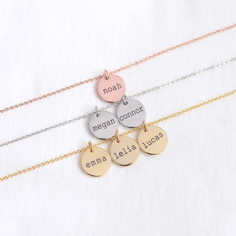 Op de afbeelding: Vijf gouden kettingen met ronde hangers, elk gegraveerd met een andere naam: Emma, Lelia, Lucas, Megan en Connor. De bovenste ketting heeft een ros&eacute;gouden ketting en een hanger gegraveerd met de naam Noah.