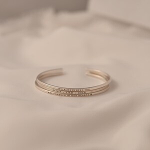 May include: Three silver-colored cuff bracelets stacked on a white surface. Each bracelet has an engraved message. The top bracelet reads "IT'S ALWAYS BEEN YOU." The second bracelet reads "OLIVIA &bull; LIAM &bull; NOAH." The bottom bracelet reads "MAX FOREVER IN OUR HEARTS" with a paw print.