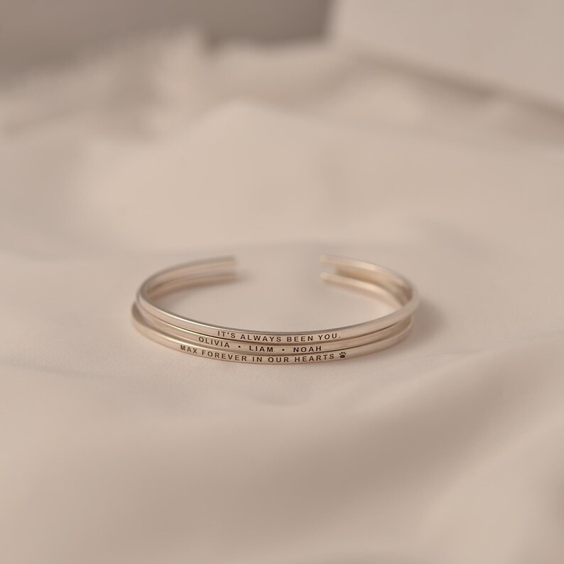 May include: Three silver-colored cuff bracelets stacked on a white surface. Each bracelet has an engraved message. The top bracelet reads "IT'S ALWAYS BEEN YOU." The second bracelet reads "OLIVIA &bull; LIAM &bull; NOAH." The bottom bracelet reads "MAX FOREVER IN OUR HEARTS" with a paw print.