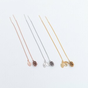 May include: Four pairs of long, delicate chain earrings with a small pine cone and leaf charm at the end. The earrings are available in silver, rose gold, and gold.
