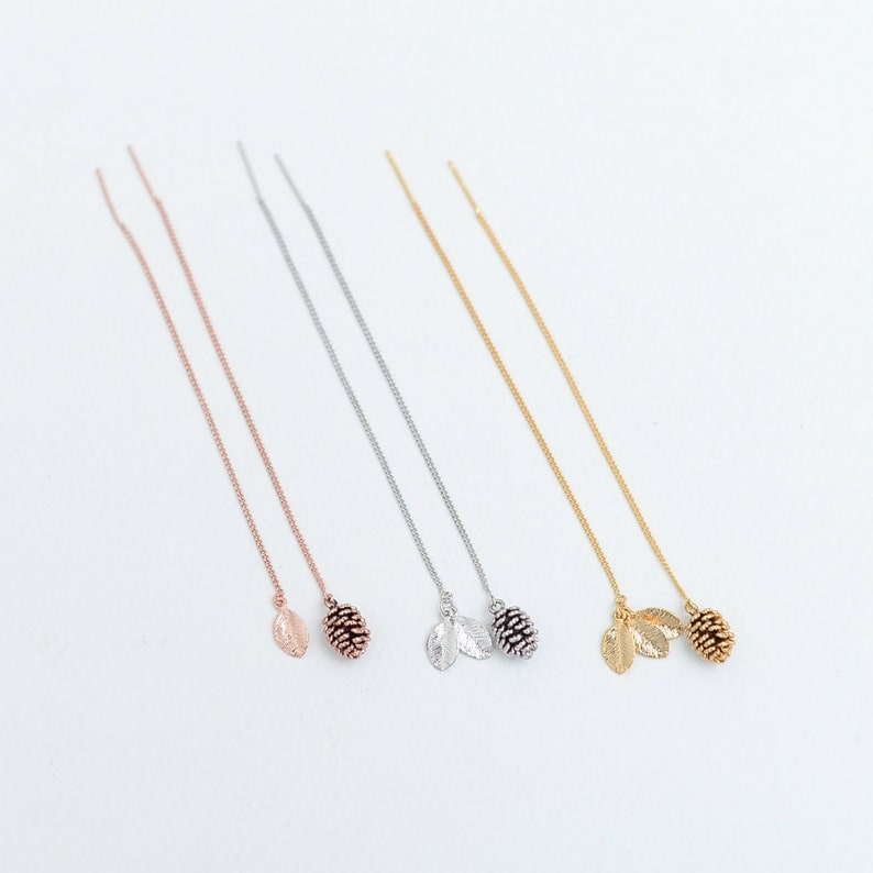 May include: Four pairs of long, delicate chain earrings with a small pine cone and leaf charm at the end. The earrings are available in silver, rose gold, and gold.