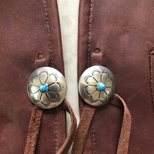 May include: Two silver and turquoise flower-shaped buttons with leather straps attached to a brown leather vest.