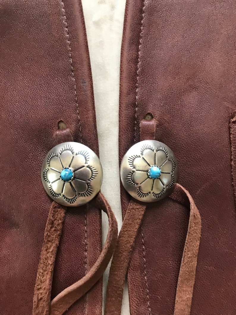 May include: Two silver and turquoise flower-shaped buttons with leather straps attached to a brown leather vest.