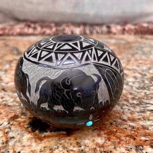 May include: Black ceramic pot with a carved design of a buffalo and geometric patterns. The pot is decorated with turquoise stones.