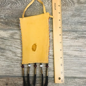 Puede incluir: Una peque&ntilde;a bolsa de cuero con cuentas, con flecos y una correa de cuero. La bolsa est&aacute; decorada con cuentas de colores en un patr&oacute;n geom&eacute;trico. La bolsa mide aproximadamente 15 cm de largo y 7,5 cm de ancho.