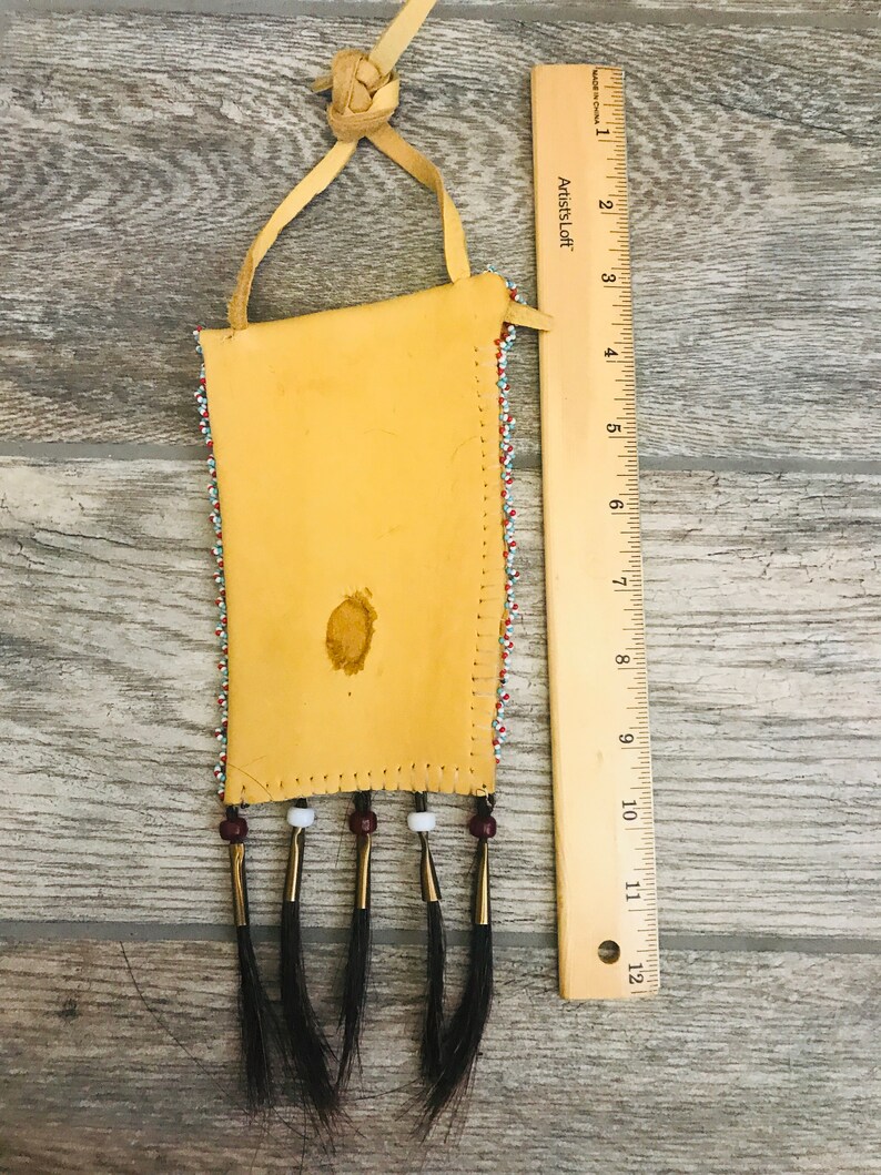 Puede incluir: Una peque&ntilde;a bolsa de cuero con cuentas, con flecos y una correa de cuero. La bolsa est&aacute; decorada con cuentas de colores en un patr&oacute;n geom&eacute;trico. La bolsa mide aproximadamente 15 cm de largo y 7,5 cm de ancho.