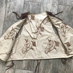 May include: A brown leather vest with a cream-colored cotton lining. The lining features a repeating pattern of brown horse heads.