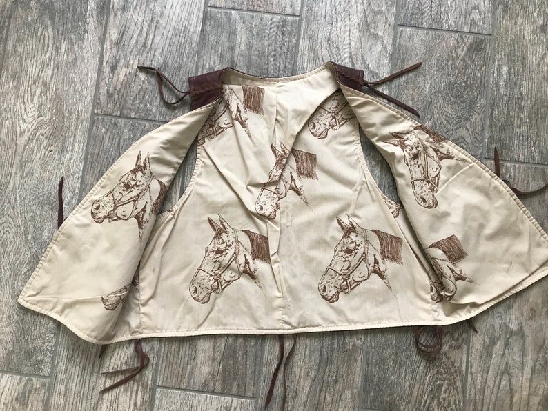 May include: A brown leather vest with a cream-colored cotton lining. The lining features a repeating pattern of brown horse heads.