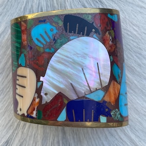 May include: A wide cuff bracelet featuring a brass frame inlaid with a mosaic of colorful stones and shell. The design includes several bear-shaped silhouettes crafted from iridescent white shell, dark blue stone, and light blue turquoise.