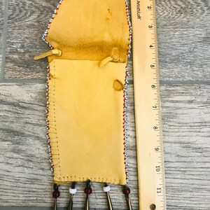 Puede incluir: Una bolsa de cuero con cuentas y flecos con una regla para la escala. La bolsa est&aacute; decorada con cuentas rojas, blancas y azules. La bolsa mide aproximadamente 30 cm de largo.