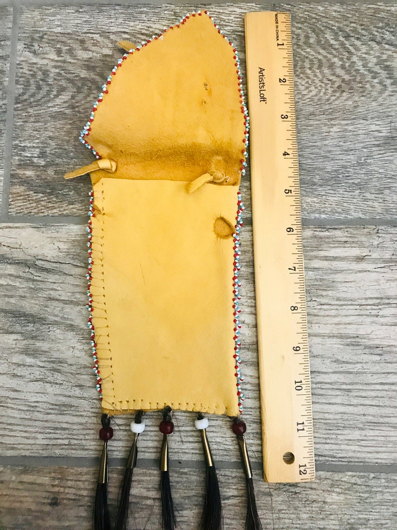 Puede incluir: Una bolsa de cuero con cuentas y flecos con una regla para la escala. La bolsa est&aacute; decorada con cuentas rojas, blancas y azules. La bolsa mide aproximadamente 30 cm de largo.