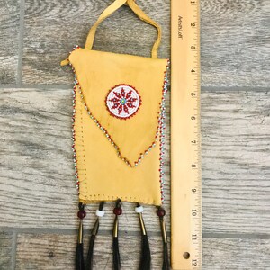 Puede incluir: Una peque&ntilde;a bolsa de cuero marr&oacute;n con un dise&ntilde;o de cuentas y flecos. La bolsa tiene un dise&ntilde;o de flor de cuentas en el centro y est&aacute; decorada con cuentas rojas, blancas y azules. La bolsa mide aproximadamente 18 cm de alto.